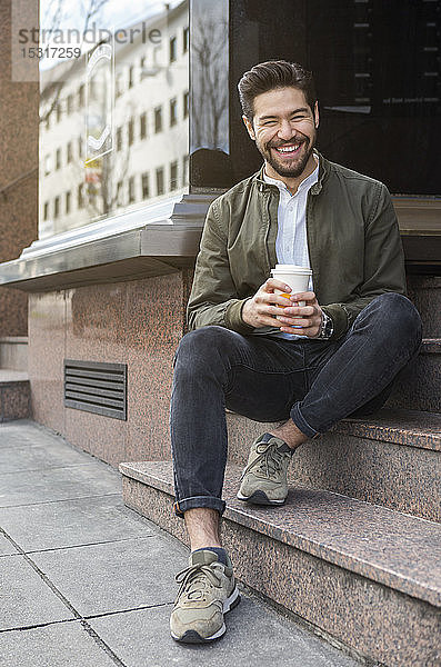 Porträt eines lachenden jungen Mannes  der mit Kaffee zum Mitnehmen auf der Treppe sitzt