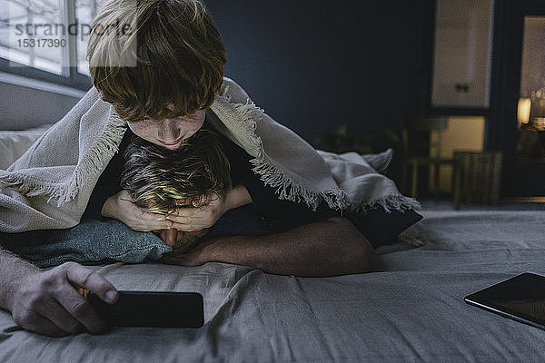 Vater und Sohn liegen zusammen unter einer Decke und benutzen ein Smartphone