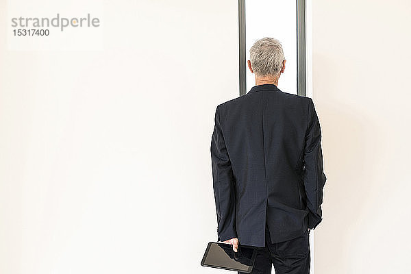 Rückansicht eines leitenden Geschäftsmannes mit Tablette  der durch ein Fenster schaut