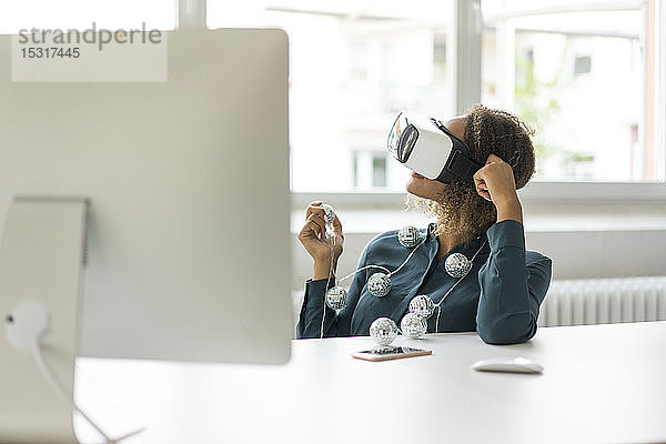 Junge Frau sitzt am Schreibtisch mit Lichterkette und benutzt eine Virtual-Reality-Brille