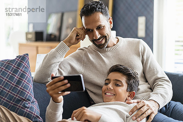 Lächelnder Vater und Sohn mit Smartphone auf der Couch im Wohnzimmer
