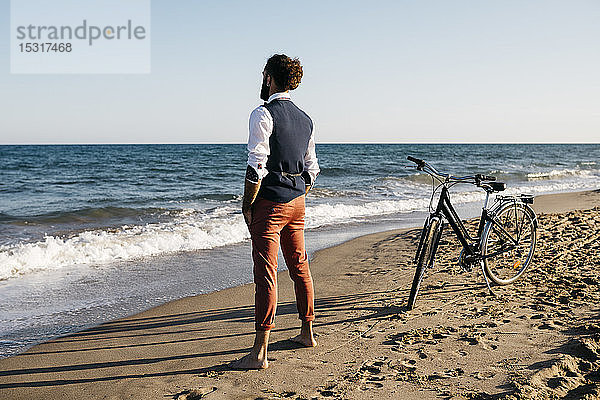 Gut gekleideter Mann mit seinem Fahrrad am Strand stehend