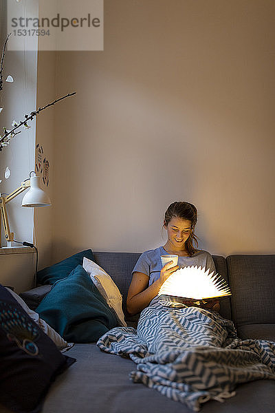 Junge Frau liest zu Hause auf dem Sofa ein illuminiertes Buch