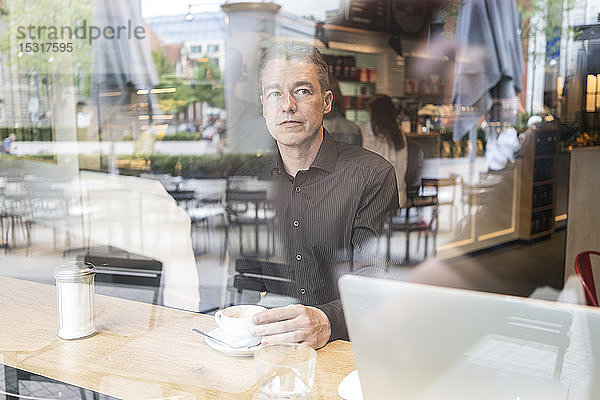 Mann sitzt in einem Café hinter einer Fensterscheibe und trinkt Kaffee mit einem Laptop