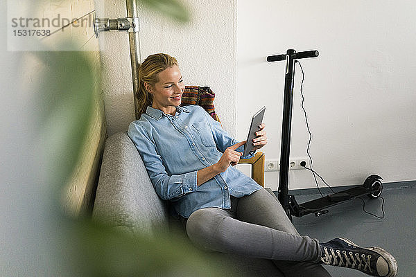 Junge Frau sitzt in einem Café auf der Couch und benutzt ein digitales Tablet  im Hintergrund lädt ein E-Scooter auf