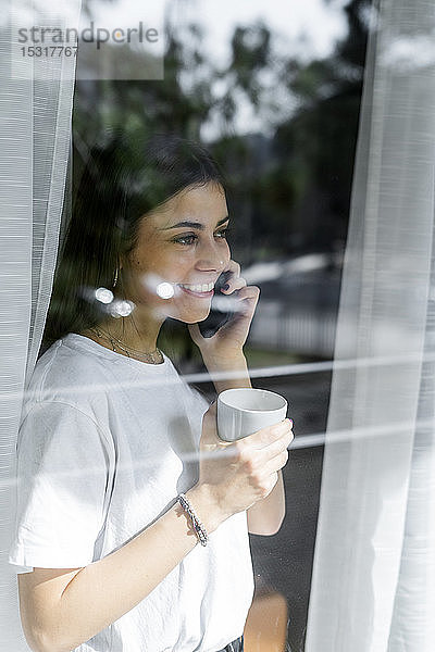 Lächelnde junge Frau am Handy hinter der Fensterscheibe