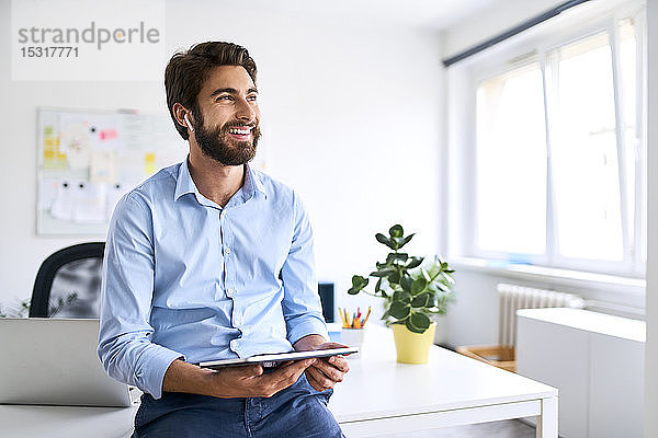 Lächelnder Geschäftsmann mit Kopfhörern  der ein digitales Tablet in seinem Büro benutzt