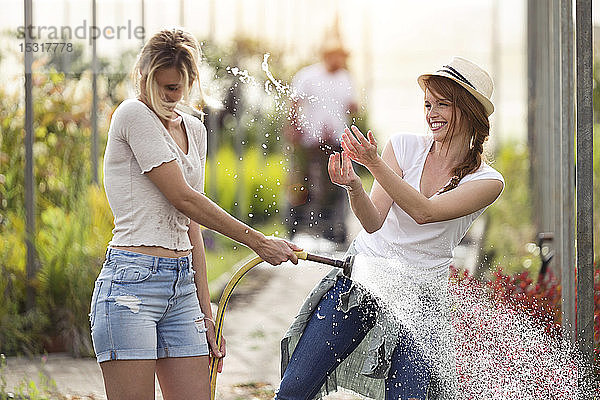Zwei glückliche junge Frauen amüsieren sich im Gewächshaus beim Gießen der Blumen mit dem Schlauch