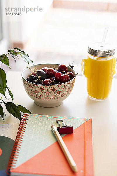 Orangen-Smoothie und Kirschschale auf Tisch mit Notizbuch und Bleistift
