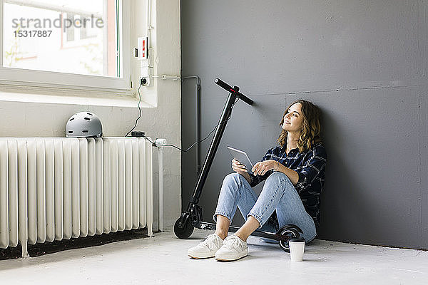 Junge Frau mit E-Scooter auf dem Boden sitzend  mit digitalem Tablet