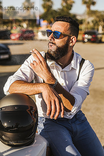 Porträt eines bärtigen Motorradfahrers mit verspiegelter Sonnenbrille  der sich auf seinen Helm stützt und raucht