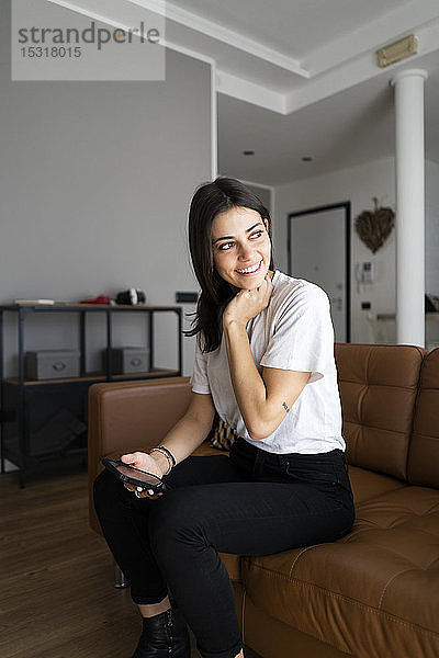 Lächelnde junge Frau auf der Couch zu Hause mit Handy