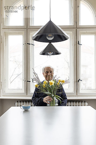 Lächelnder älterer Mann mit Blumenstrauss sitzt zu Hause am Tisch