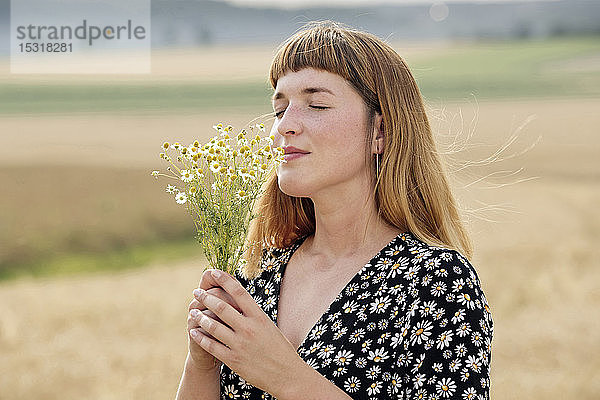 Lächelnde junge Frau mit geschlossenen Augen riecht in der Natur nach Kamillenblütensträußchen