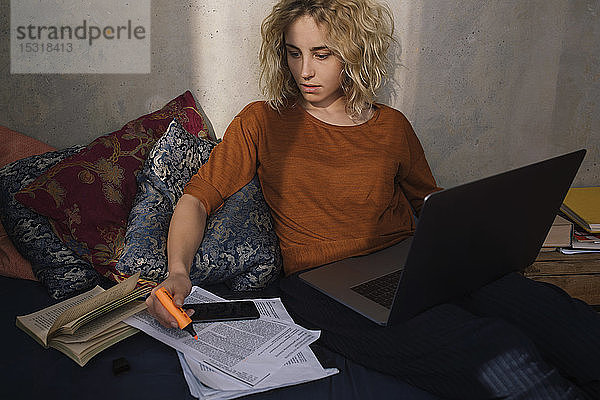 Porträt einer blonden Studentin  die mit Laptop auf dem Bett sitzt und arbeitet