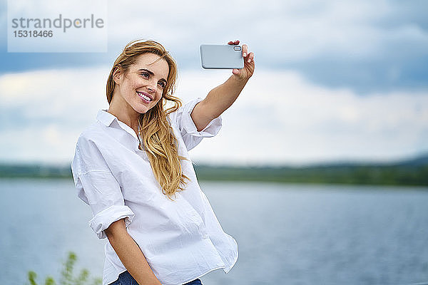 Glückliche Frau  die mit einem Smartphone am Seeufer Selbsthilfe betreibt