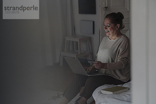 Glückliche ältere Frau sitzt zu Hause im Bett und benutzt Kreditkarte und Laptop