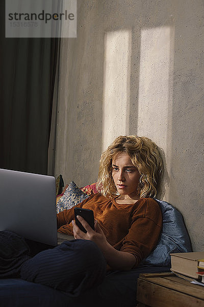 Porträt einer blonden jungen Frau  die mit Handy und Laptop auf dem Bett liegt