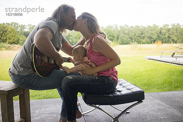Mann mit Gitarre küsst Frau mit auf dem Schoß sitzendem Mädchen