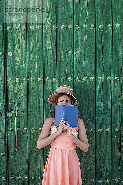 Porträt einer jungen Frau  die im Sommer mit einem Buch vor einer grünen Holztür steht