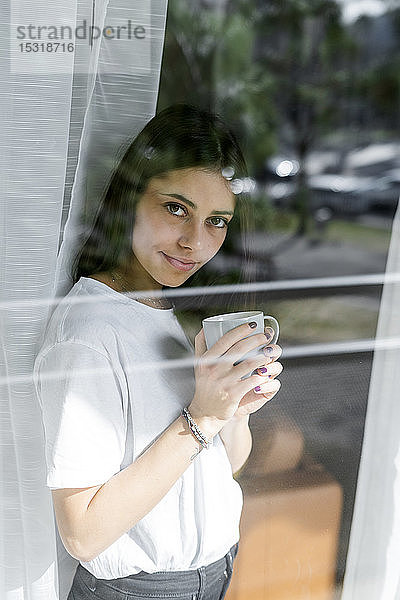 Porträt einer jungen Frau mit einer Tasse Kaffee hinter einer Fensterscheibe