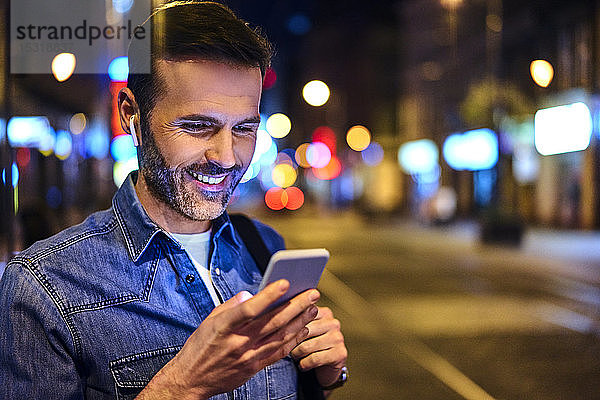 Lächelnder Mann mit drahtlosen Kopfhörern  der nachts in der Stadt ein Smartphone benutzt