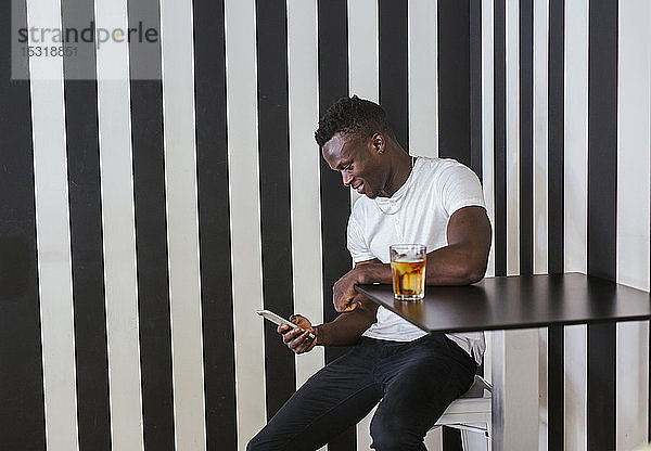 Junger Mann sitzt mit einem Getränk am Tisch und benutzt sein Handy