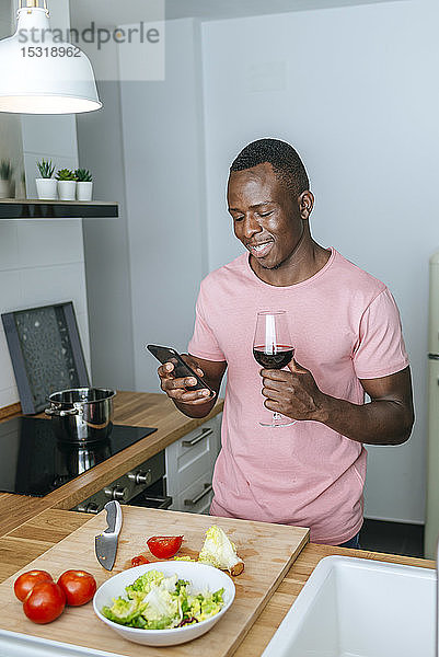 Lächelnder junger Mann mit Weinglas  der während der Salatzubereitung sein Handy benutzt