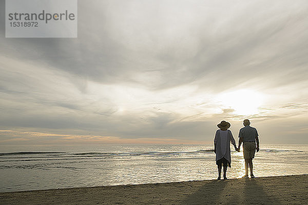 Rückenansicht eines älteren Ehepaares  das Hand in Hand am Strand steht und den Sonnenuntergang beobachtet  Liepaja  Lettland