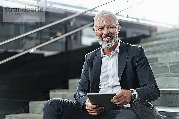Porträt eines lächelnden  reifen Geschäftsmannes auf einer Treppe sitzend mit Tablette