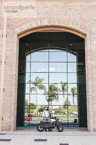 Mann mit Motorrad vor Glasfassade stehend