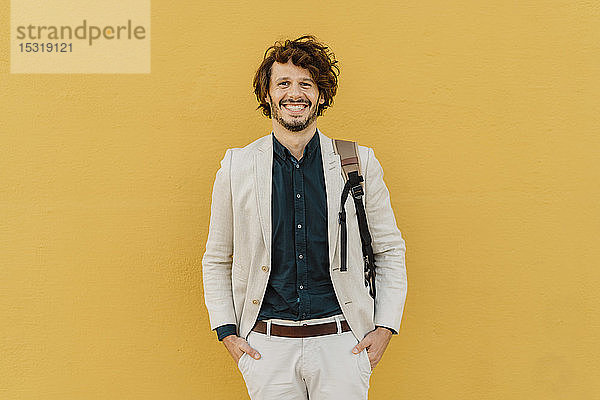 Porträt eines lachenden Geschäftsmannes mit Rucksack vor gelber Wand stehend