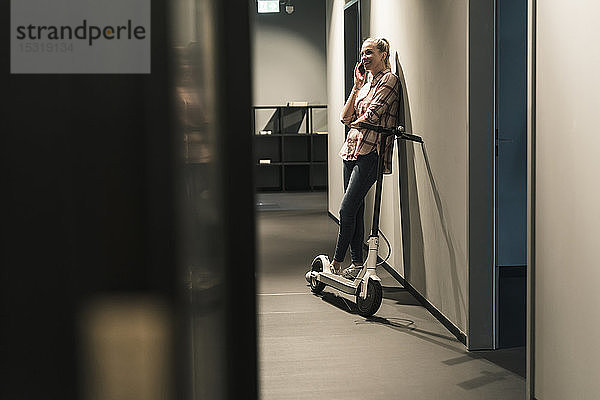 Lächelnde Geschäftsfrau mit E-Scooter  die im Büro mit dem Handy telefoniert