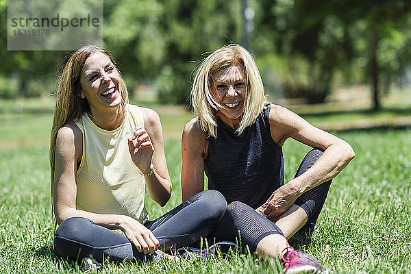 Glückliche sportliche reife Frau und ihre Tochter sitzen auf einer Wiese in einem Park