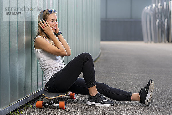 Lächelnde junge Frau sitzt auf einem Longboard und hört Musik mit Kopfhörern