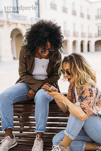 Multikulturelle glückliche Frauen  die sich unterhalten und ein Smartphone benutzen  auf einer Bank sitzend