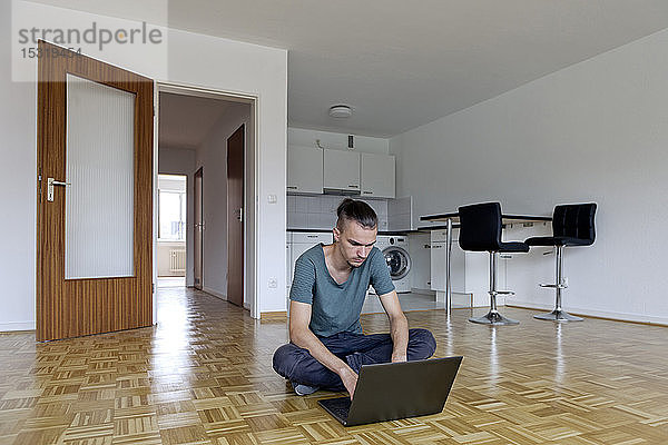 Junger Mann sitzt in einer leeren Wohnung und benutzt seinen Laptop