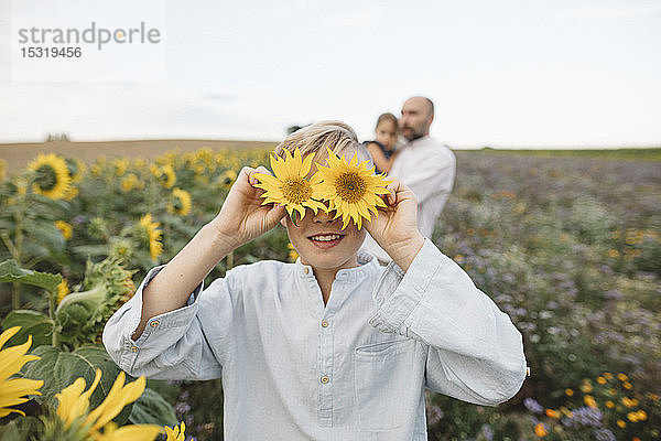 Verspielter Junge  der seine Augen mit Sonnenblumen auf einem Feld mit Familie im Hintergrund bedeckt