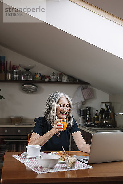Porträt einer lachenden älteren Frau mit Laptop am Frühstückstisch