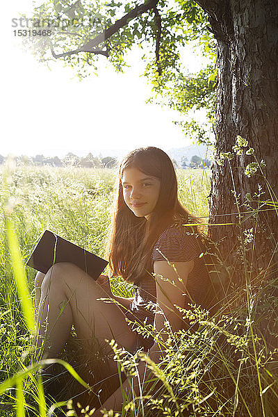 Porträt eines lächelnden Mädchens mit Buch  das an einen Baumstamm auf einer Wiese gelehnt ist