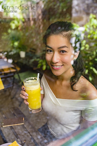 Porträt einer lächelnden jungen Frau  die ein gesundes Getränk hinter einer Glasscheibe hält