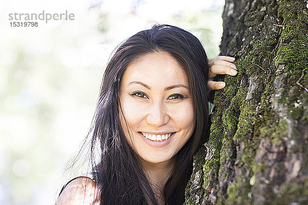 Porträt einer jungen lächelnden Frau  die sich an einen Baumstamm lehnt