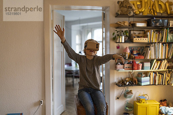 Junge sitzt auf einem Bock in seinem Zimmer zu Hause und trägt eine VR-Pappbrille