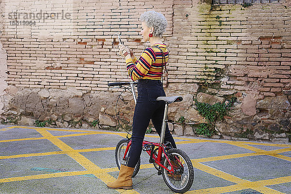 Modische reife Frau mit Fahrrad  die Ohrhörer und Smartphone benutzt