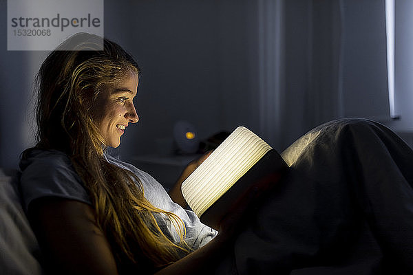 Lächelnde junge Frau liest zu Hause im Bett ein illuminiertes Buch