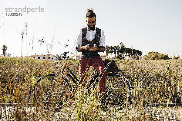 Gut gekleideter Mann mit seinem Fahrrad auf einem Holzsteg auf dem Land  der mit seinem Handy telefoniert
