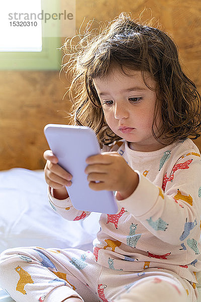 Süßes kleines Mädchen im Pyjama im Bett mit Mobiltelefon