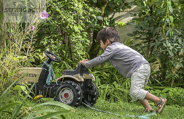 Vierjähriger Junge im Garten  der seinen Spielzeugtraktor schiebt