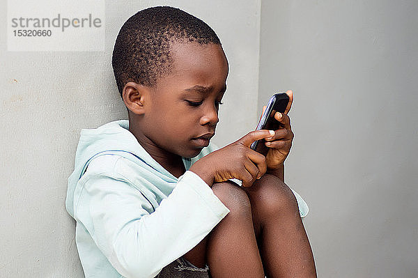 ein Kind  das an der Wand sitzt und neugierig mit einem Mobiltelefon hantiert.