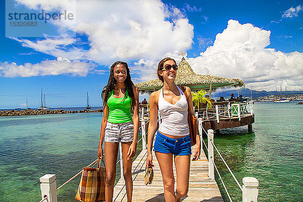 2 lächelnde Freundinnen  die auf einem Steg aus einer Bar auf Stelzen an einem karibischen Strand in einer paradiesischen Landschaft kommen.